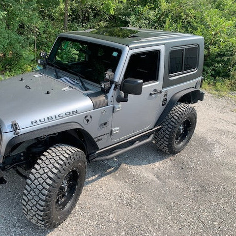 Gray Jeep Wrangler with black rims parked on a gravel surface with greenery in the background