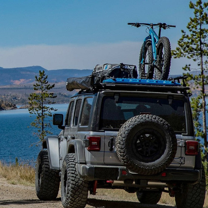Jeep with a bike on top, parked by a lake with mountains in the background