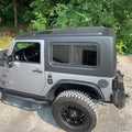 Gray Jeep Wrangler with black rims parked on a gravel surface with greenery in the background