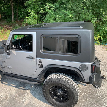 Gray Jeep Wrangler with black rims parked on a gravel surface with greenery in the background