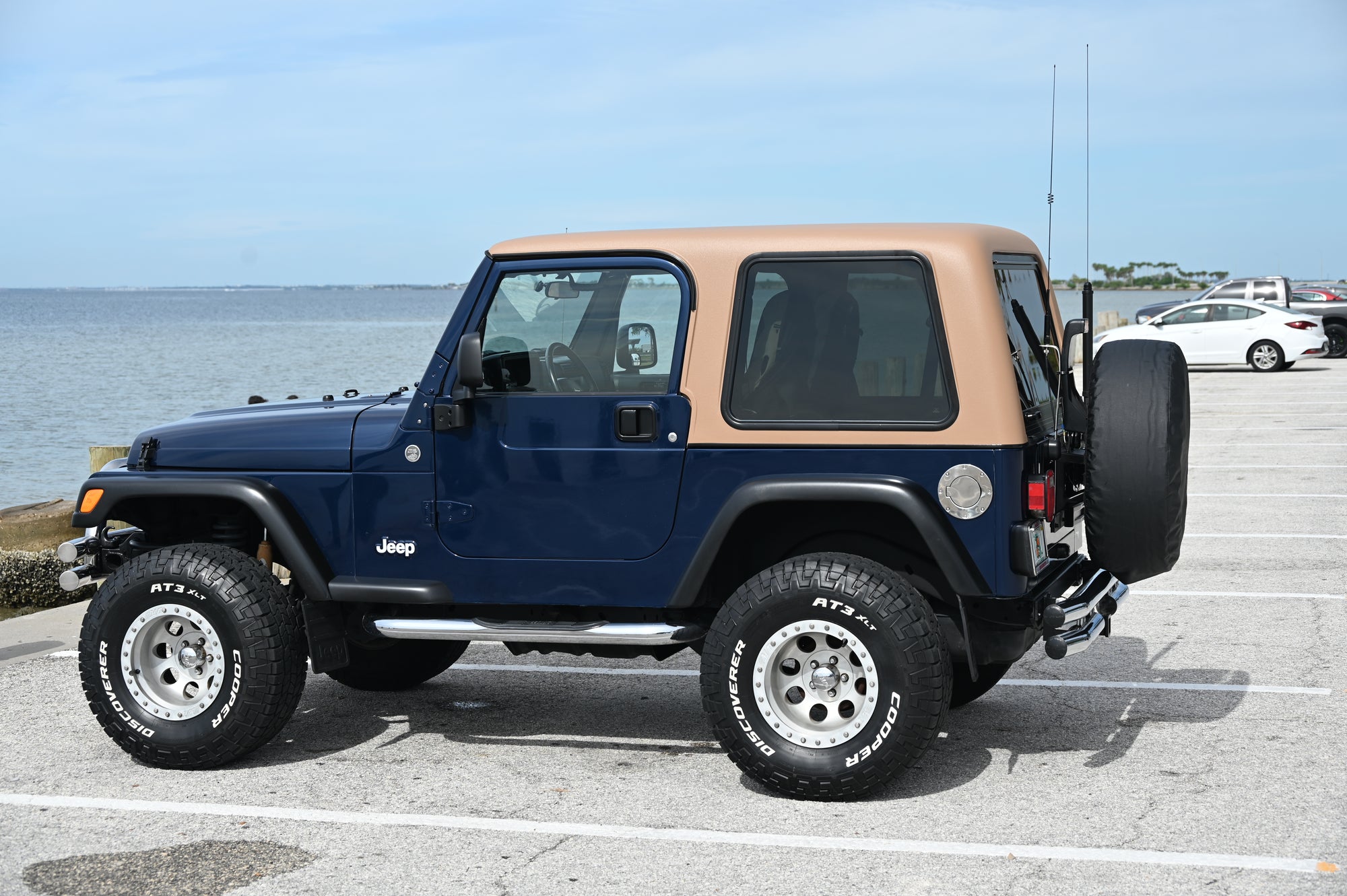 Blue Jeep Wrangler with tan roof parked in a lot near water