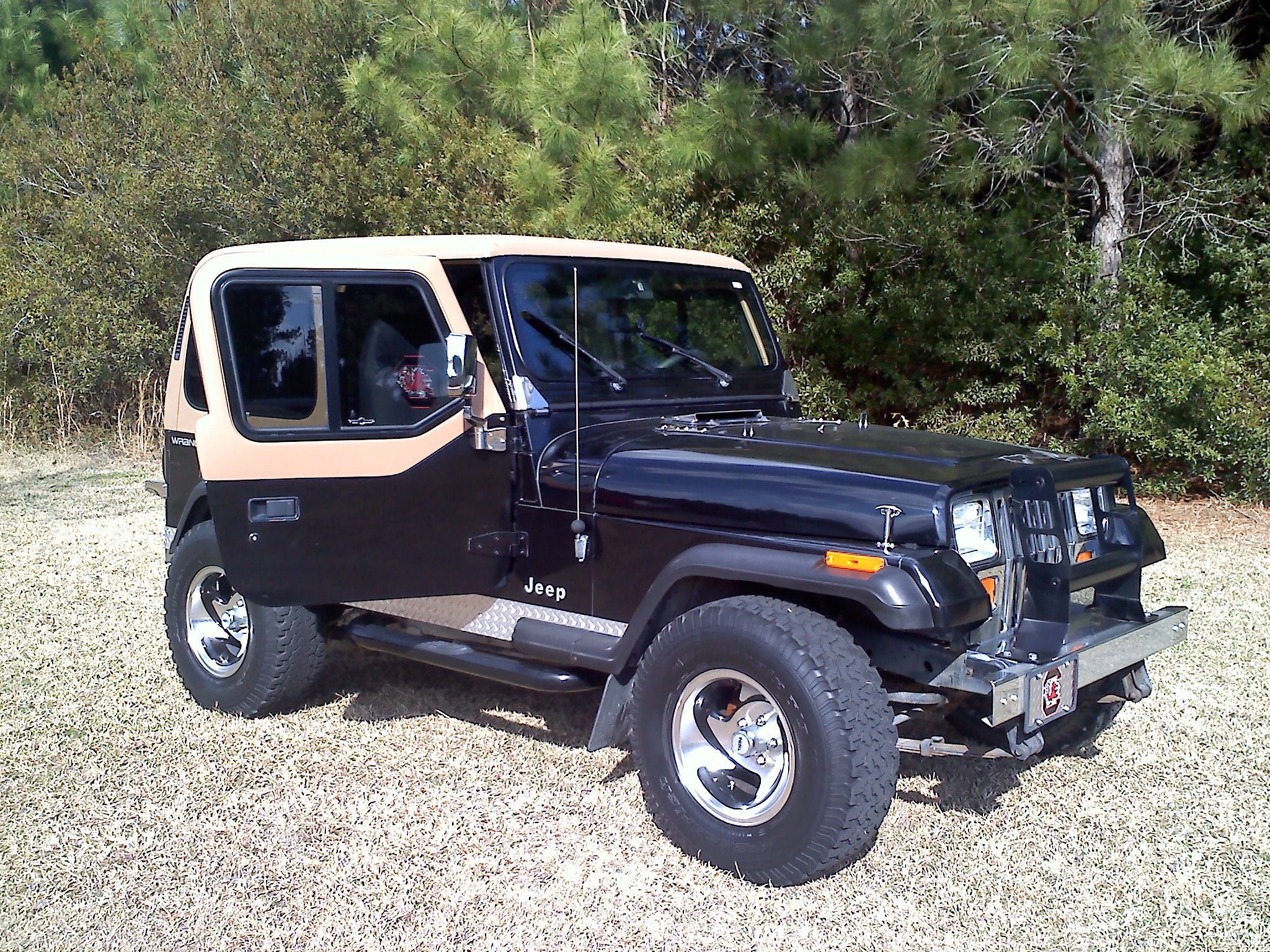 Black Jeep Wrangler with beige door on a gravel surface with greenery in the backgroundslider_item_pGKzHL