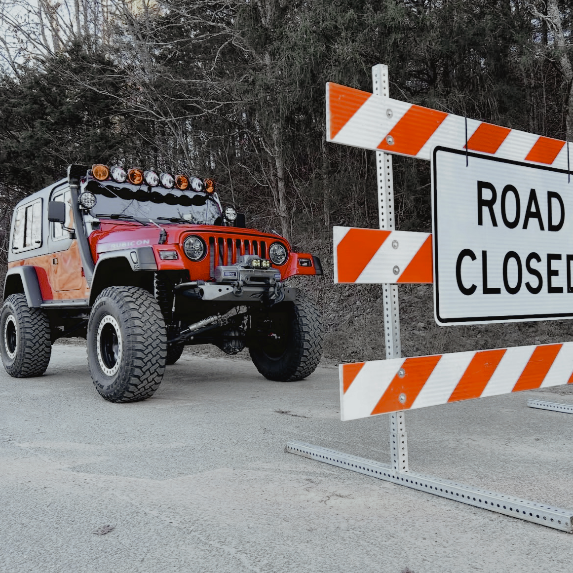 Jeep with large tires near a 'Road Closed' sign in a forested area