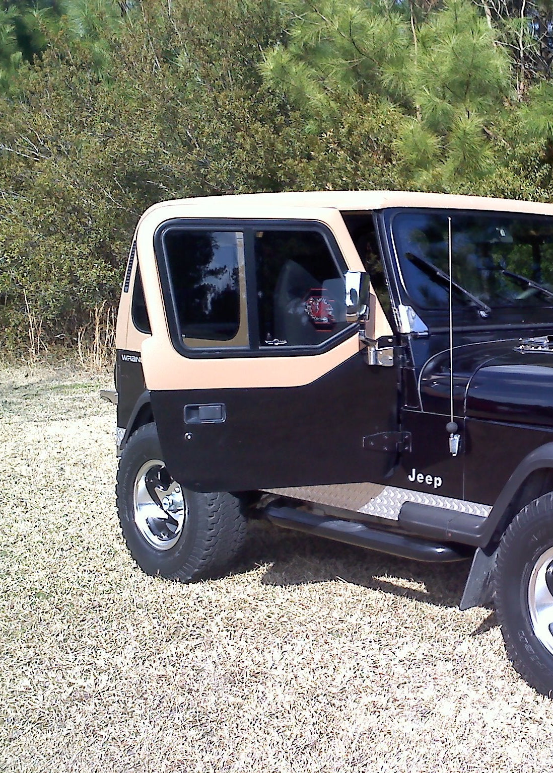 Black Jeep with a beige roof parked on a gravel surface with greenery in the background